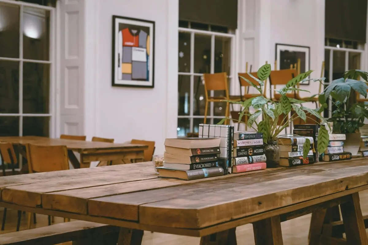 A wooden table with stacks of books and potted plants in a well-lit room with framed art on the walls and wooden chairs near the windows, creating an ambiance that promotes sober living.