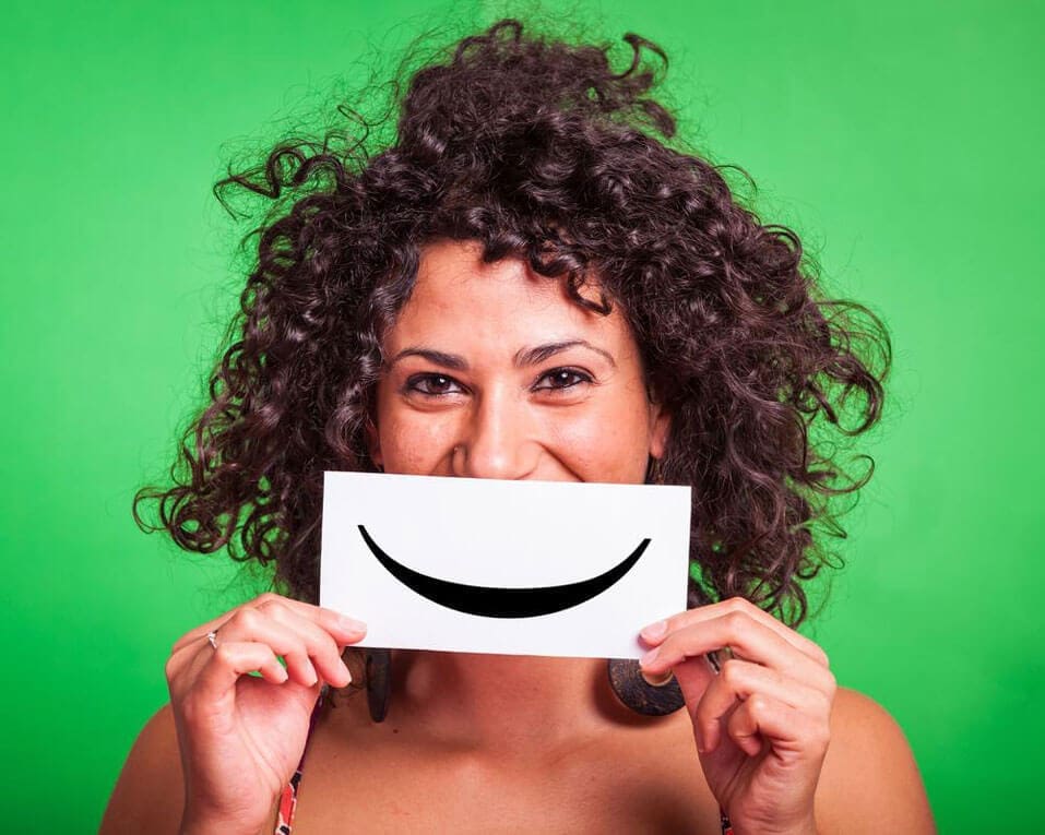 A woman is standing against a green background while holding up a card with a smile painted on it