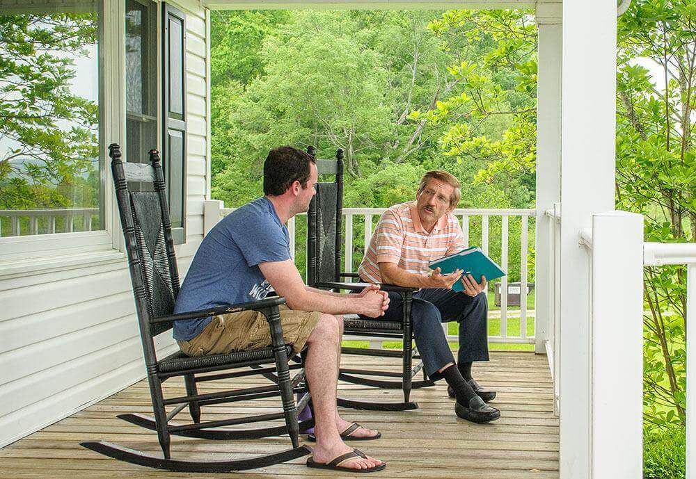 Two men sitting in rocking chairs on a porch at a drug detox center.