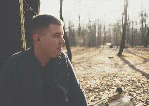A guy is sitting on a log in what looks to be a forest.