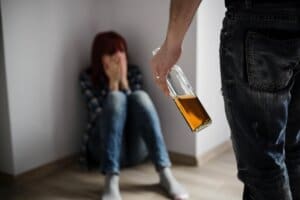 A man is holding a bottle of alcohol in front of a woman at a men's only rehab center.